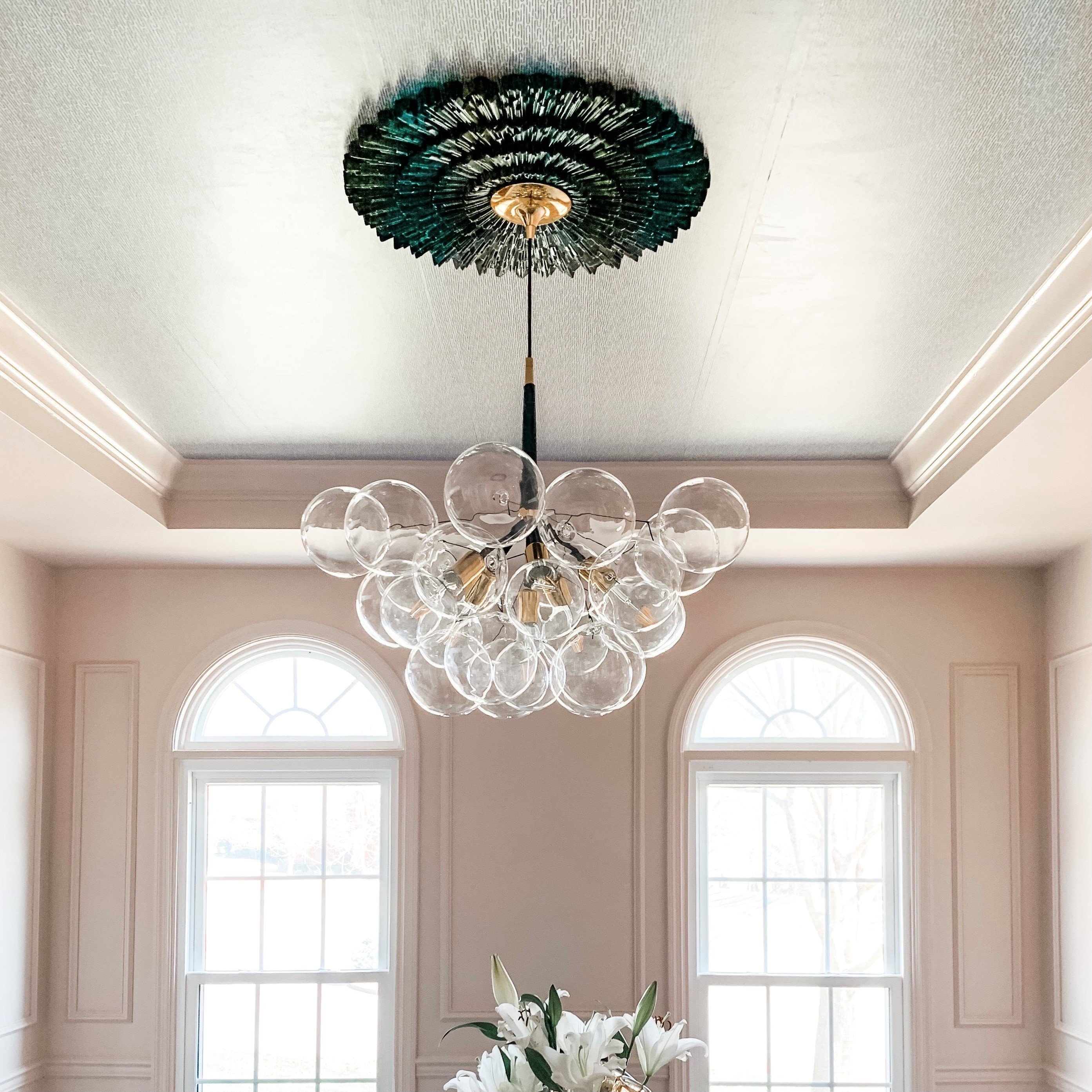 A stylish ceiling featuring an ornate medallion and a contemporary chandelier with glass orbs, illuminating a bright room with large, arched windows. Soft pastel walls complement the elegant design, creating a sophisticated and inviting atmosphere.
